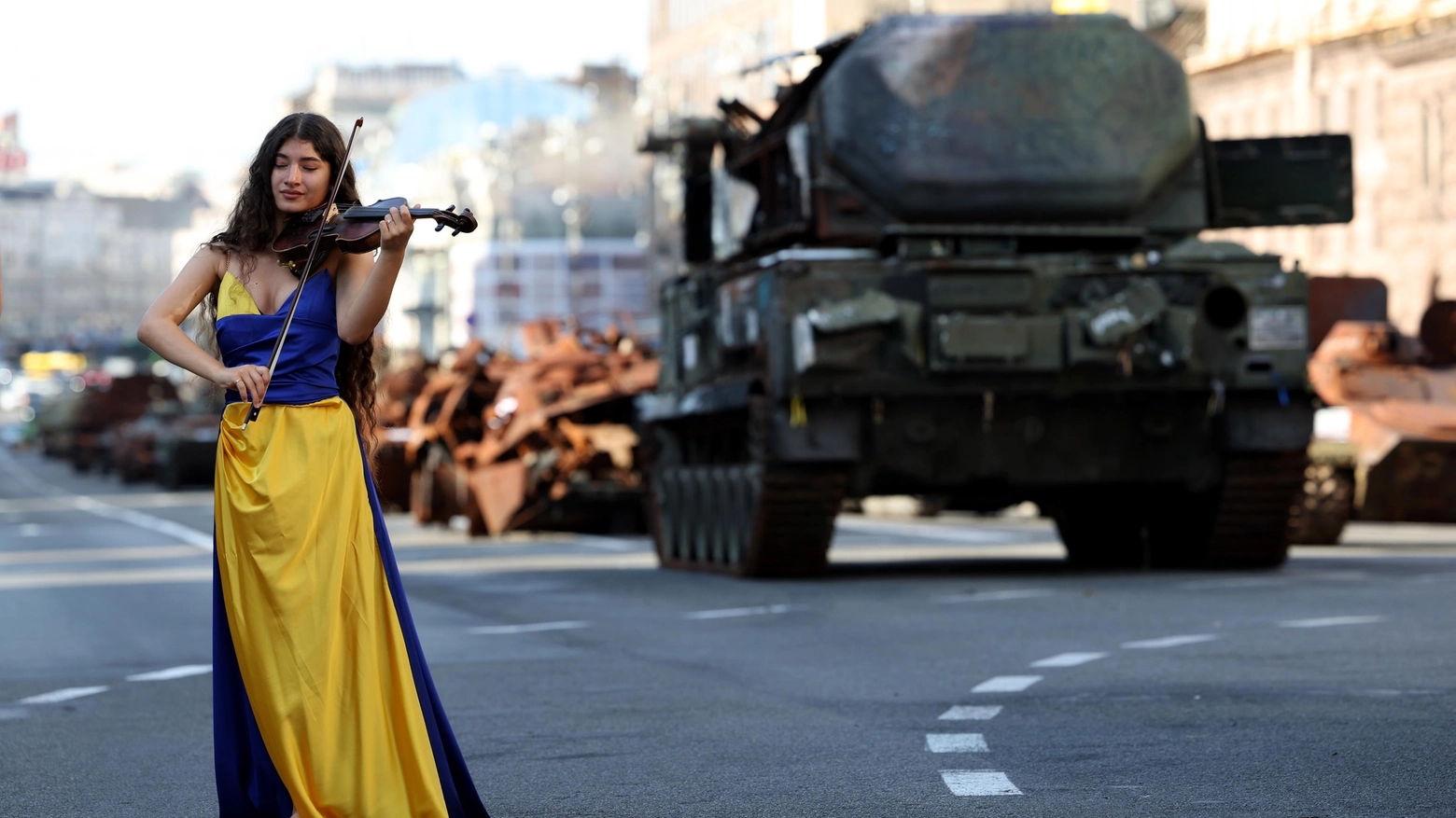 Una donna vestita con i colori dell'Ucraina suona il violino mentre cammina in una strada di Kiev fra i mezzi militari russi sequestrati