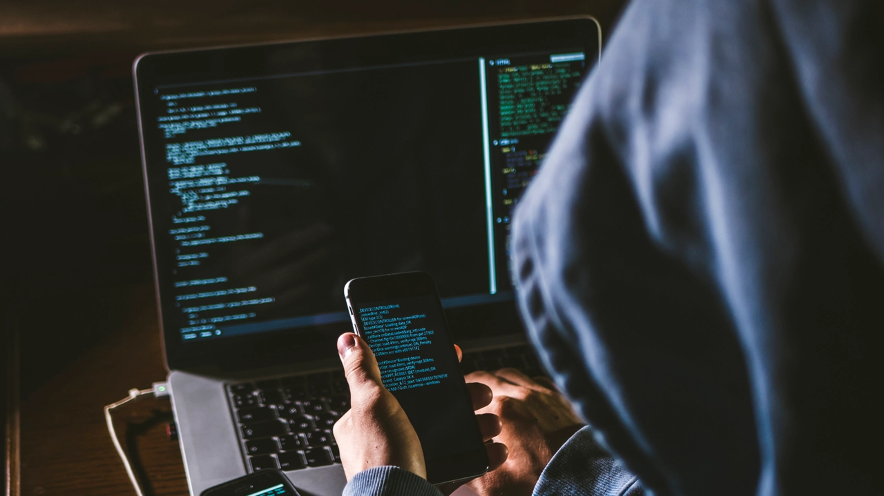 anonymous person in the hood sitting in front of computer working with laptop and mobile phone