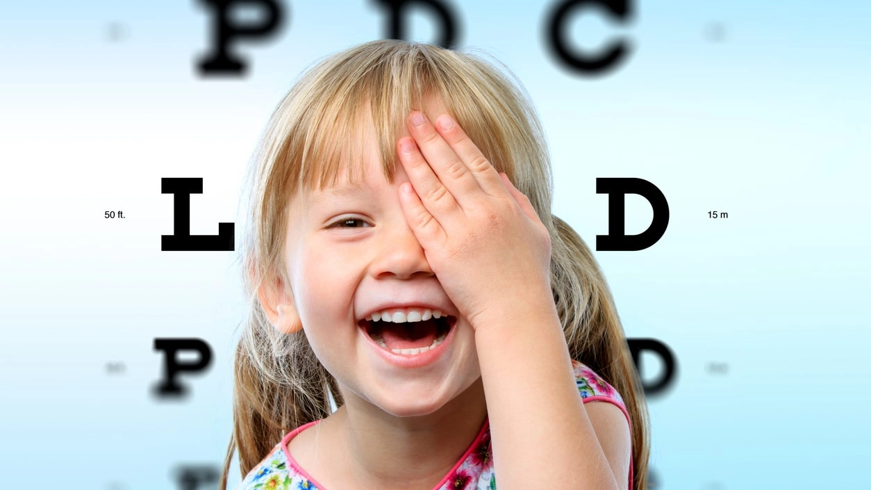 Close up face portrait of happy girl having fun at vision test.Conceptual image with girl closing one eye with hand and block letter eye chart in background.