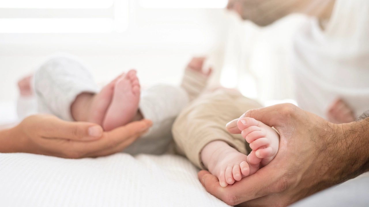 A new mother and father each hold the feet of their newborn fraternal twins laying on a bed. Shot is focused on feet and hands only.