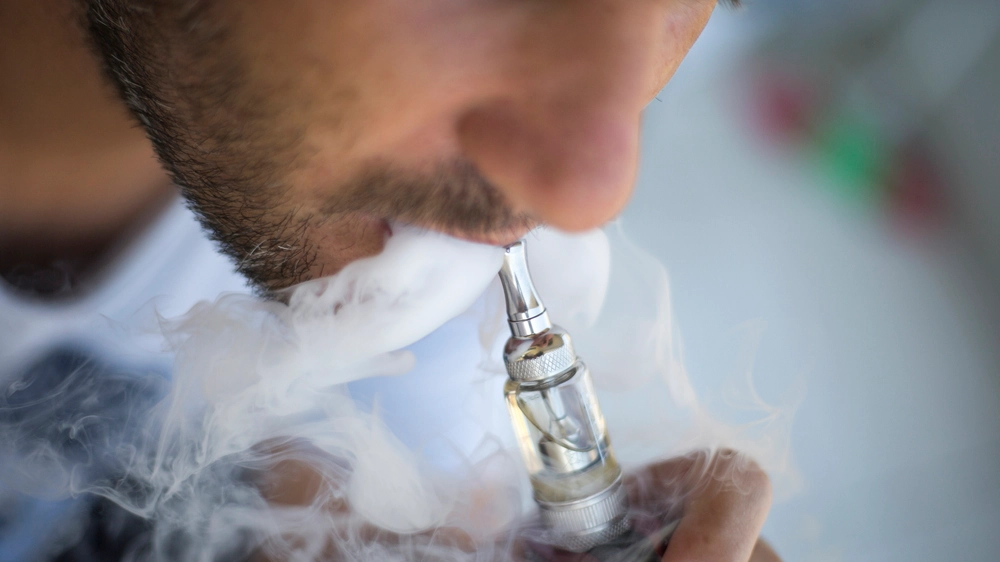 A man smokes an electronic cigarette on a gray background, blowing a stream of smoke