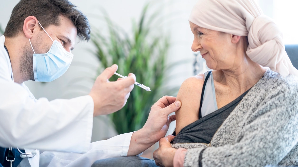 A woman wearing a headscarf allows a doctor to inject her with a vaccine. She is in her home.